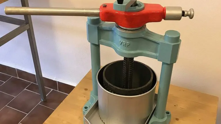 An old cast-iron screw press with a red handle bar and a metal drum, used for squeezing liquid out of soaked herb