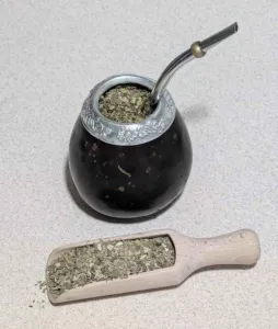 Dark calabash mate gourd with a metal bombilla straw beside a scoop of dried yerba mate leaf