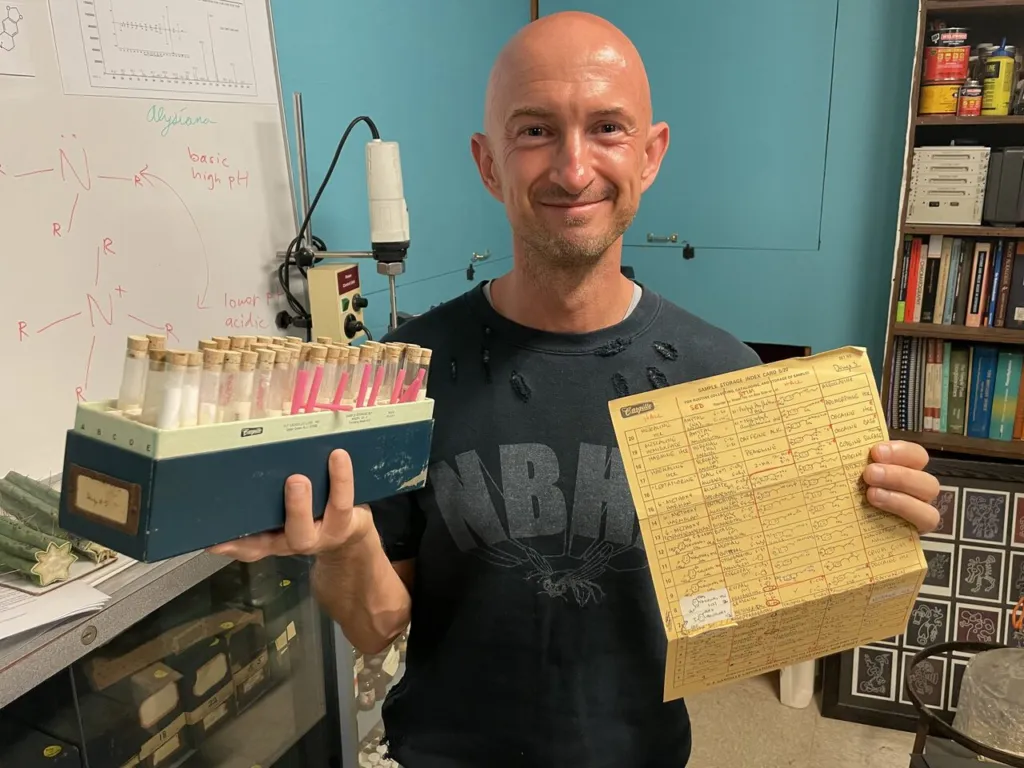 Researcher Andrew Gallimore holding a rack of sample vials and a handwritten notebook card inside Alexander Shulgin's laboratory in 2025, with structures drawn on a whiteboard behind him